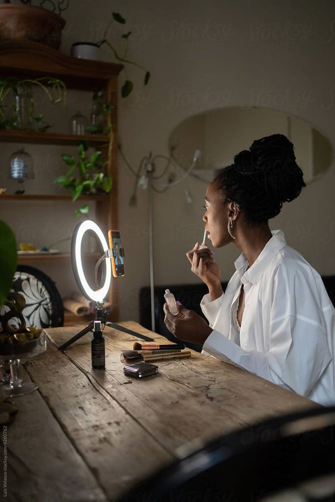 Profil of a black woman filming herself with a ringlight putting on lipstick at home on her desk for UGC.