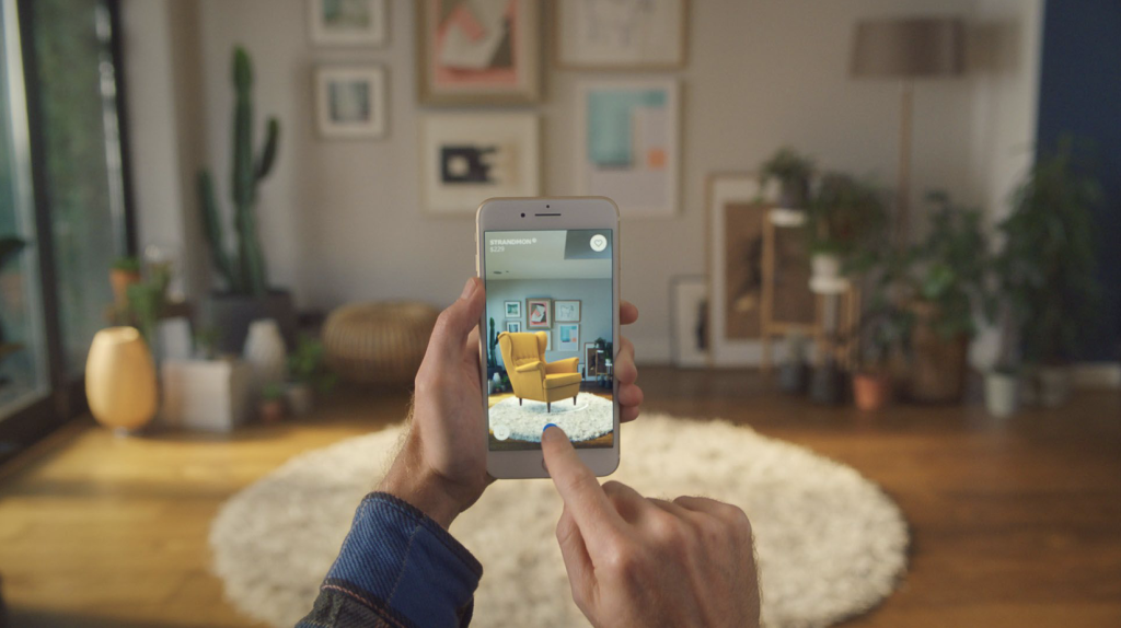 Un individu tient son téléphone devant son salon et montre le fonctionnement de l'application de réalité augmentée d'IKEA en superposant l'image d'un canapé dans l'espace.