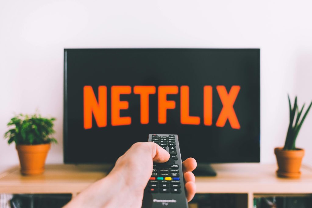 Main tenant une télécommande pointée vers un écran de télévision affichant le logo de Netflix, avec des plantes en arrière-plan sur un meuble de télévision.