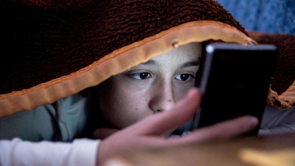 Un jeune enfant sous les couverture consulte TikTok. Cela peut être vu comme une dépendance envers l'application, potentiellement en raison de l'algorithme.
