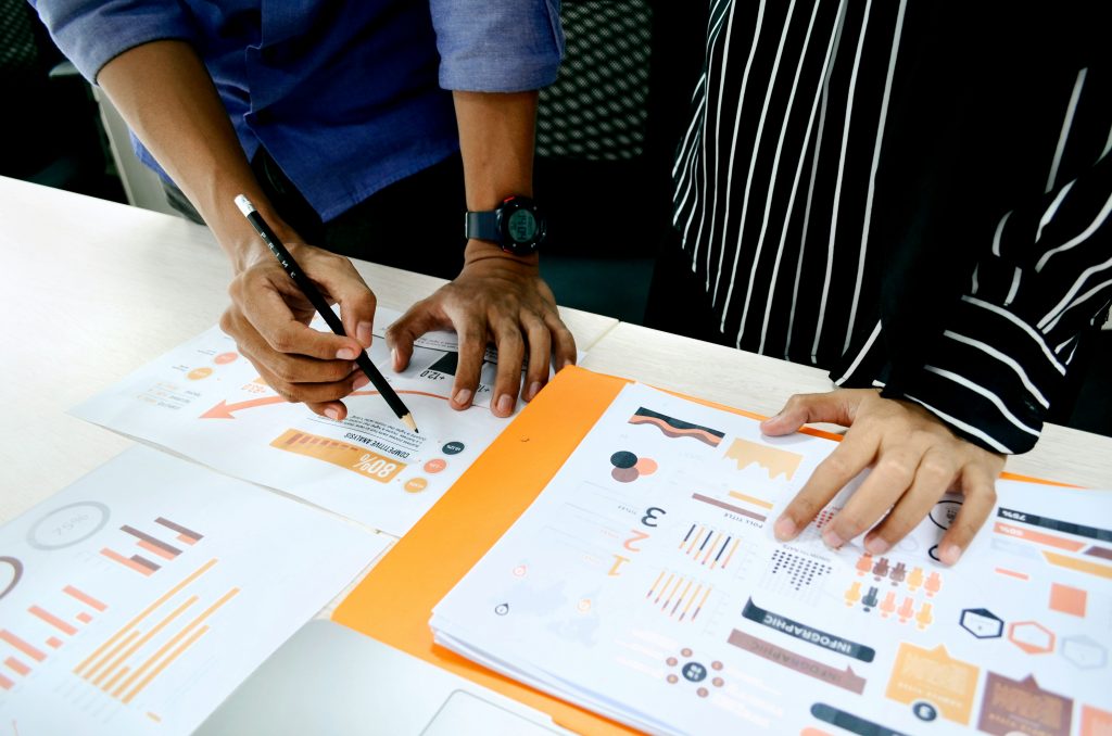 Two people analyzing papers with graphs and data. One person wearing blue shirt, second wearing black with white stripes.