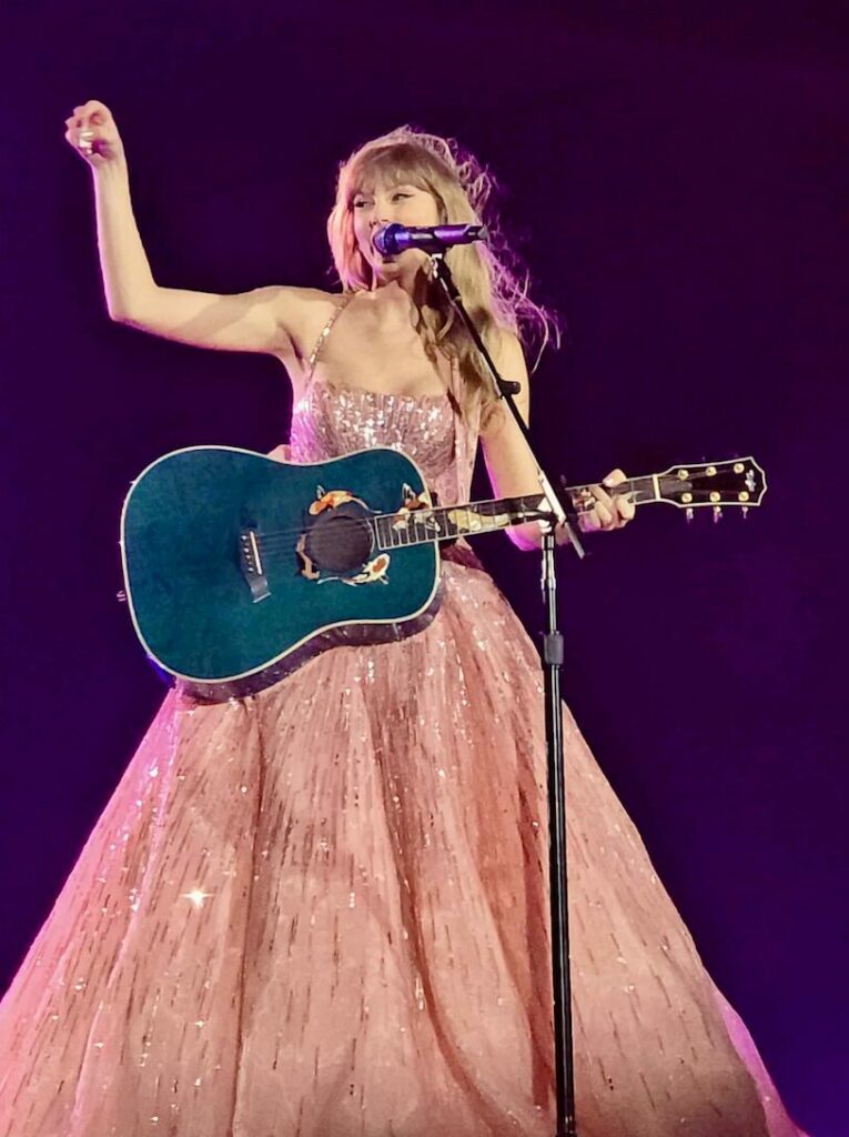 Taylor Swift performing in a sparkling pink ball gown during The Eras Tour’s Speak Now era, representing nostalgia, storytelling, and emotional connection through music, storytelling and branding. Taylor Swift in a sparkling pink ball gown, playing a blue guitar under purple stage lighting during the 'Eras Tour', symbolising her emotional connection with her audience and the final encore moment of the Speak Now era. Photo by Christopher Mao, used with permission.