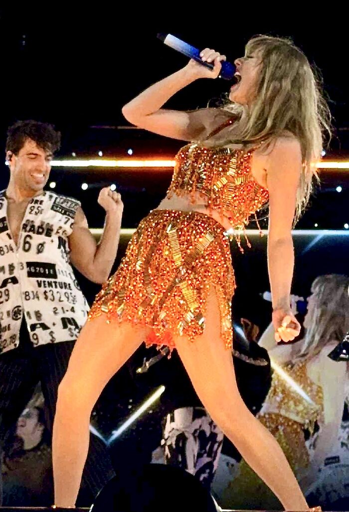 Performer in an orange sequined outfit singing powerfully on stage under bright lights during The Life of a Showgirl era, symbolising Taylor Swift’s digital marketing storytelling. Photo by Tenielle Nuske.