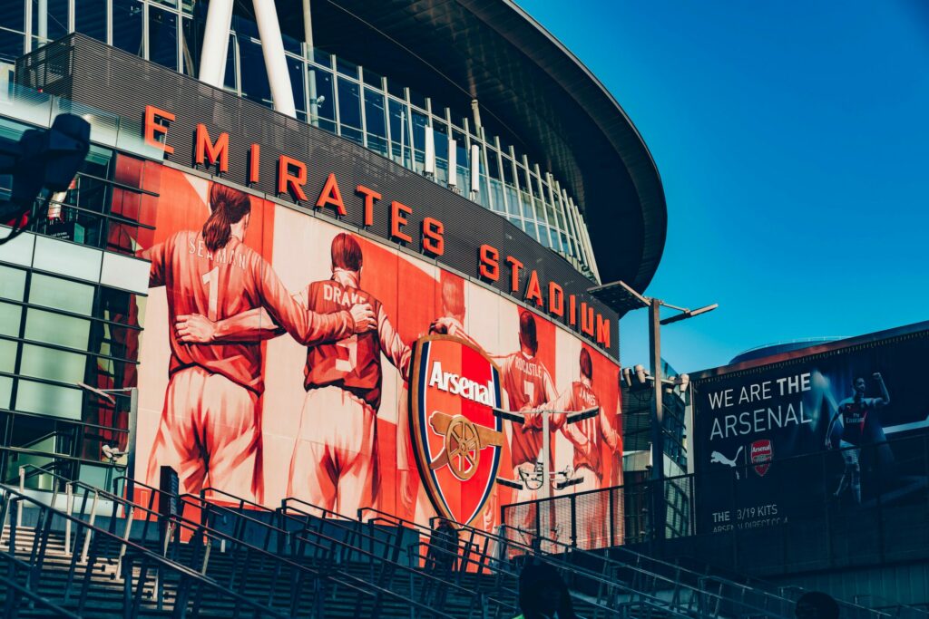 Photo of the Arsenal stadium with the name visible 'Emirates stadium', the badge seen on the stadium and a art piece of Arsenal legends surrounding the badge.