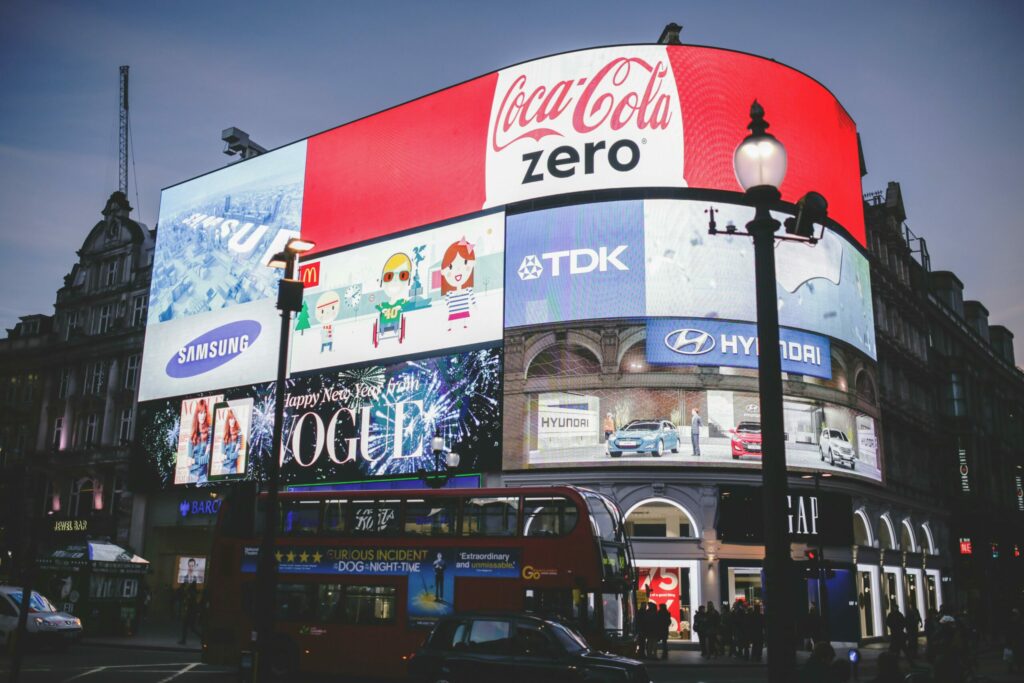 Grand écran publicitaire illuminé à Piccadilly Circus, Londres, affichant des annonces de marques comme Coca-Cola, Samsung, Hyundai et Vogue, avec un bus rouge à impériale passant au premier plan au crépuscule.