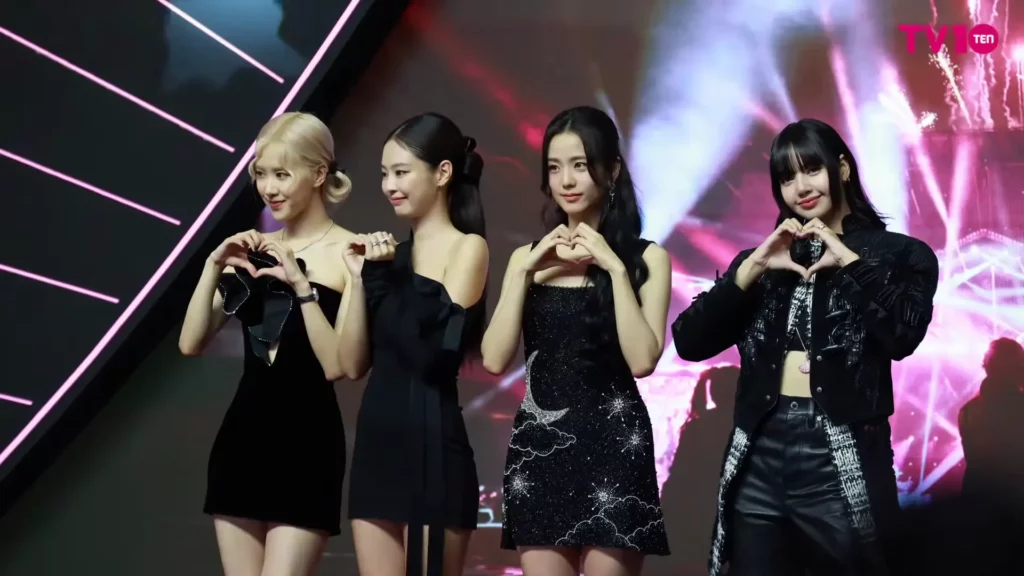 Membres groupe k-pop Blackpink sur scène lors d’un événement, cœurs avec leurs mains sur fond lumineux coloré