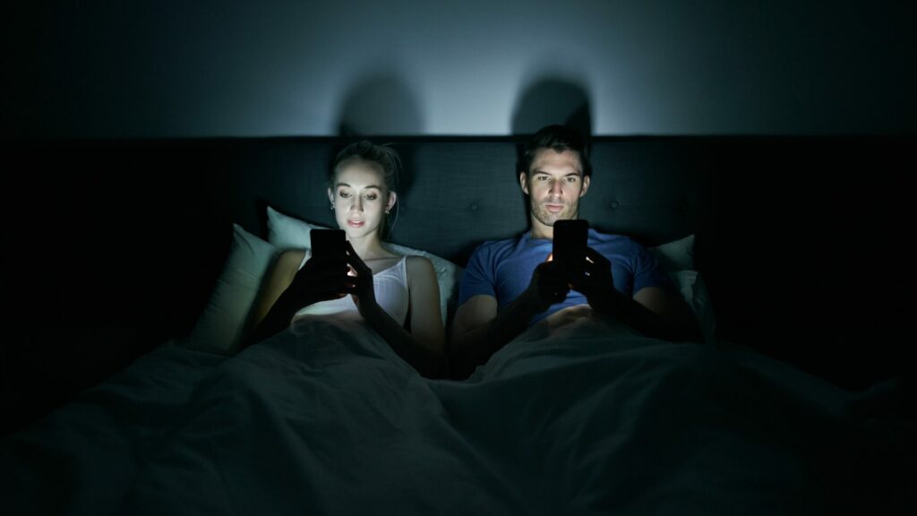 Photo d'un jeune couple qui sont sur leur téléphone dans le lit le soir.