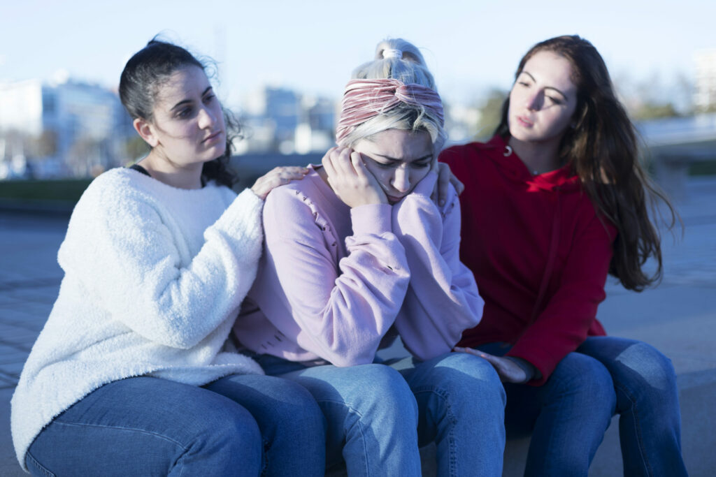 Three young women sit together on outdoor stone steps. The woman in the center appears distressed with her head in her hands, while her two friends sit on either side, offering comfort with supportive hands on her shoulder and knee.